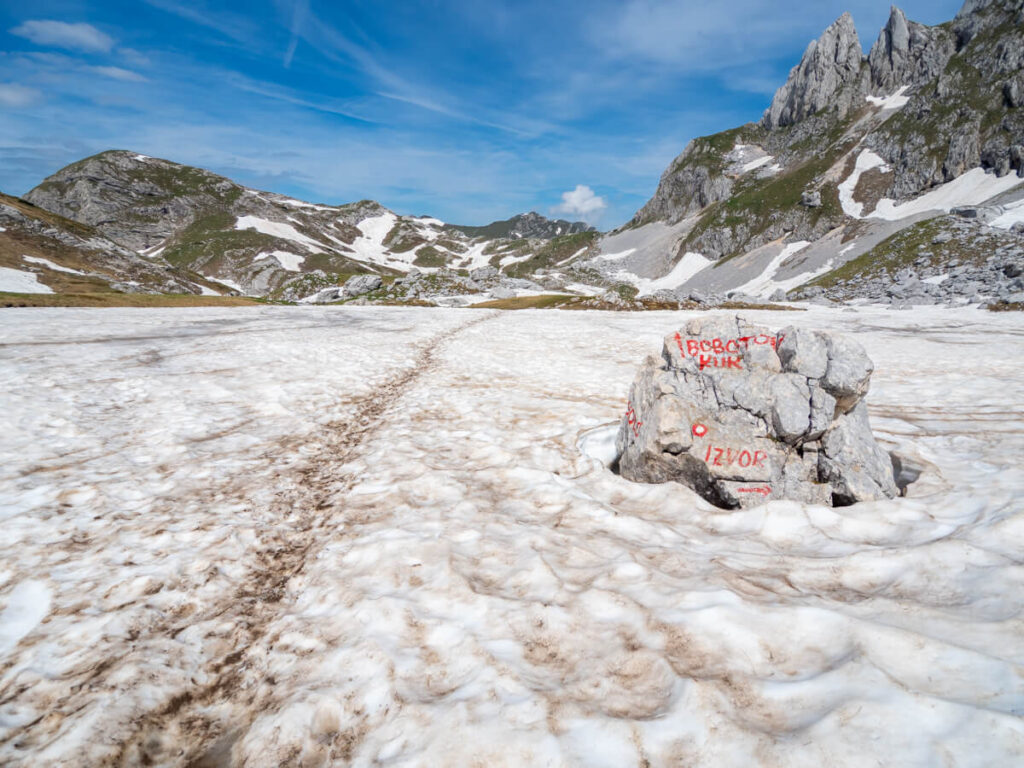 A trail of footprints leading through snow past a boulder with painted signs for Bobotov Kuk.