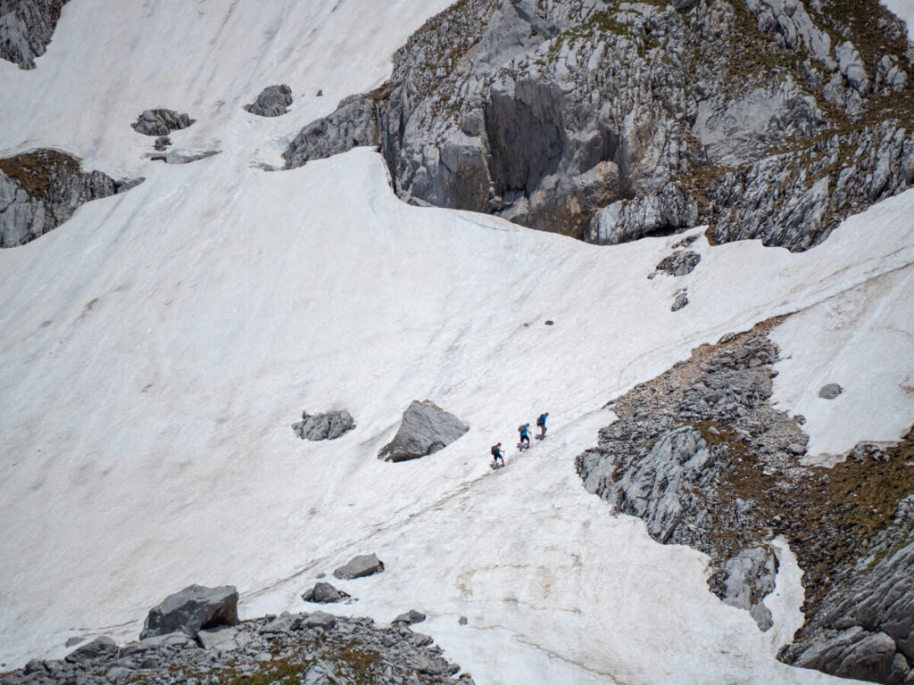 Three hikers in a line following a path of footprints through snow up the steep side of Bobotov Kuk.