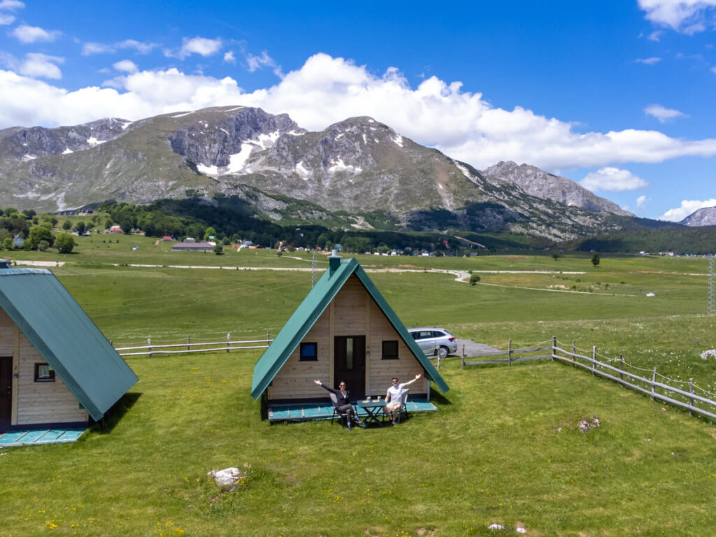 Lucy and Dan with their arms stretched wide sitting outside a wooden A-frame cabin with views of Savin Kuk and the mountains of Durmitor National Park.