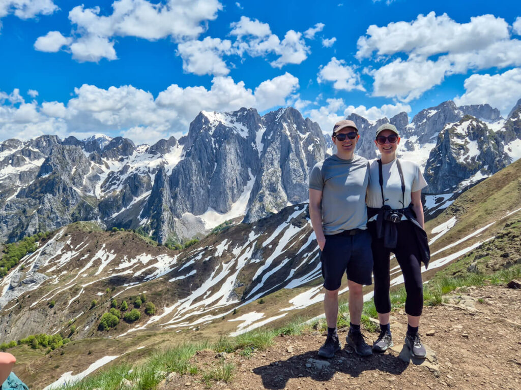 Lucy and Dan standing on Popadija in front of snowy mountains of the Karanfili massif on the Three Peaks trail in Prokletije National Park.