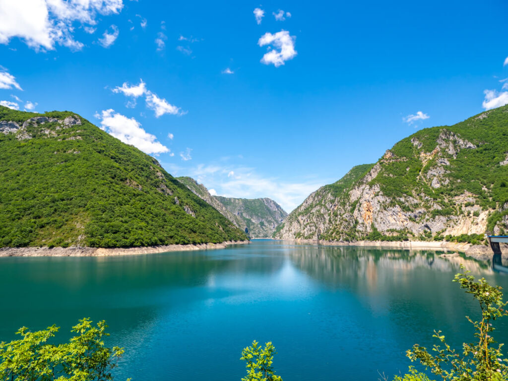 Piva Lake Valley on an almost cloudless sunny day. The water is calm and is reflecting the rocks and greenery that surround the lake.