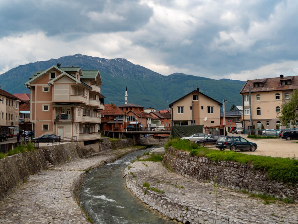 A small river winding through a town of buildings with a mosque minaret against a mountain backdrop in Plav, Montenegro.