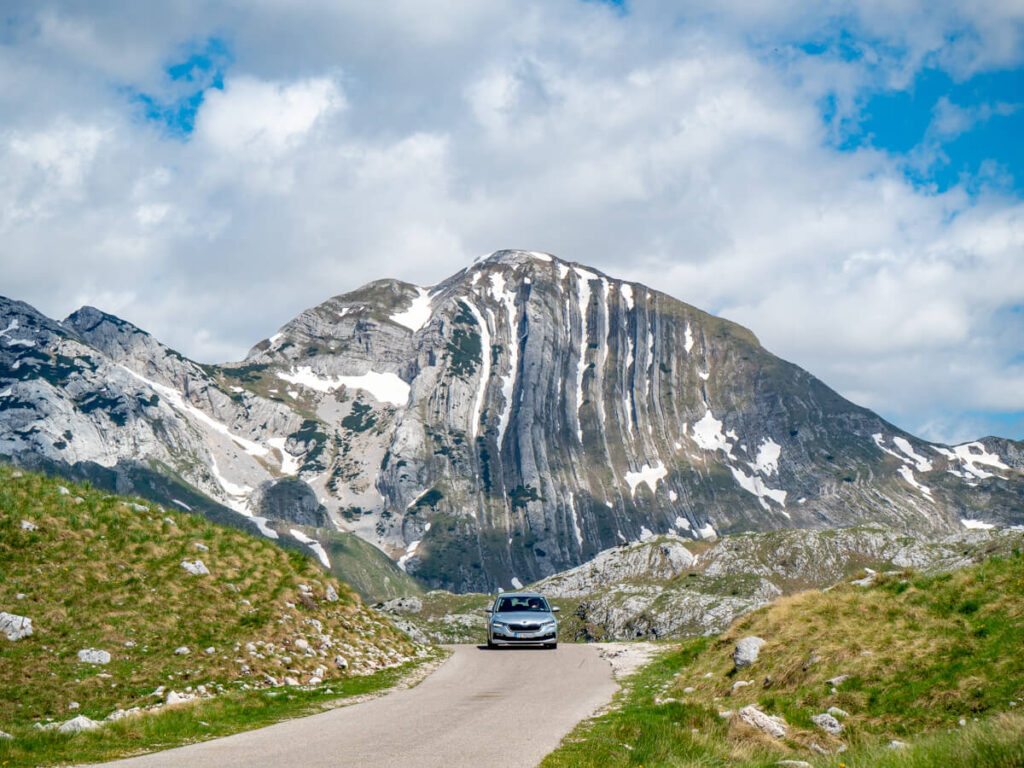 A silver car driving the Durmitor Ring Road, backed by the layers of Prutaš.