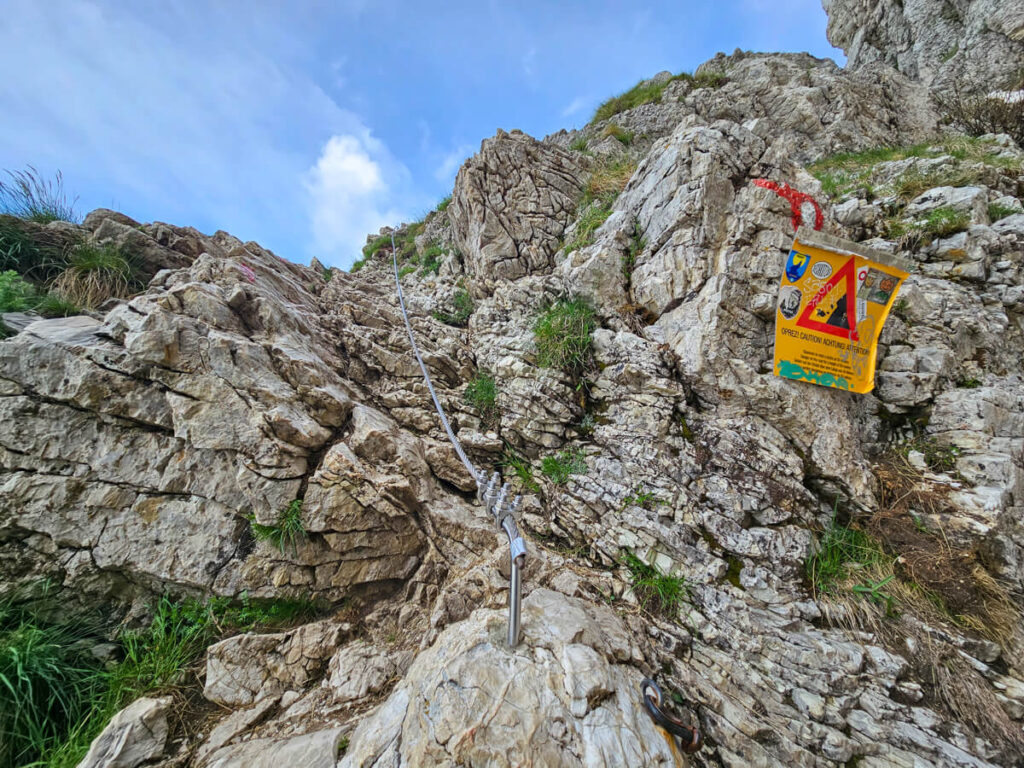 A yellow warning sign by a steel cable leading up a rockface on the Bobotov Kuk hike.