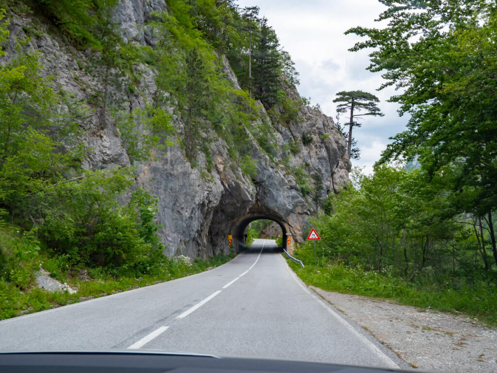 A road leading through a small tunnel carved into the rocky wall of a cliff in the mountains of Montenegro.