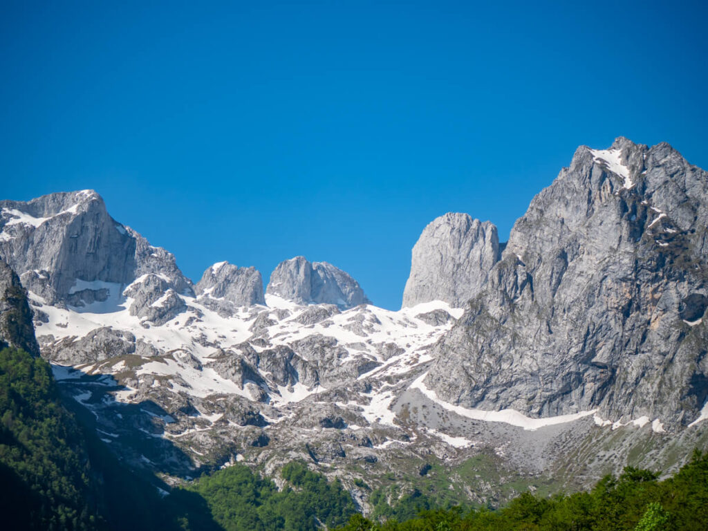 Snowy jagged limestone cliffs atop green forest in the Grebaje Valley in Prokletije National Park.