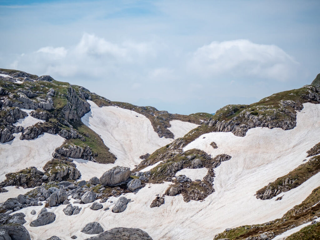 Snow spread across grey limestone boulders in a green valley on the Bobotov Kuk trail.