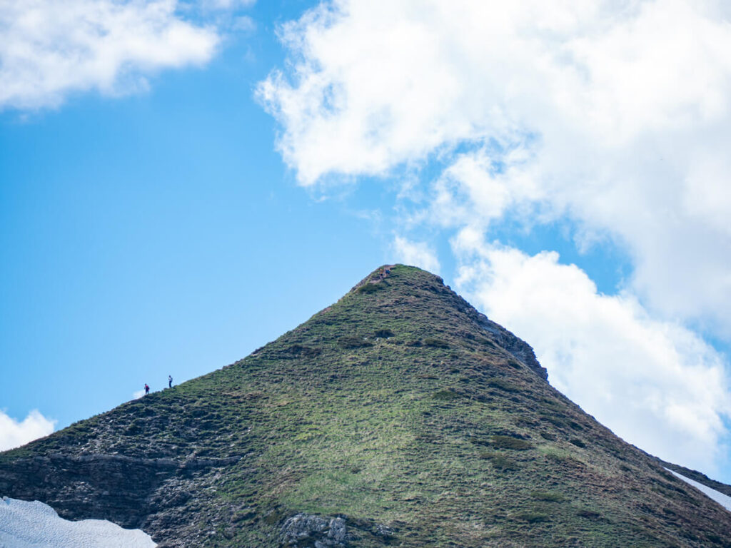 Tiny hikers on the side of the tall and steep-sided Talijanka peak strewn with snow on the Three Peaks loop in Prokletije National Park.