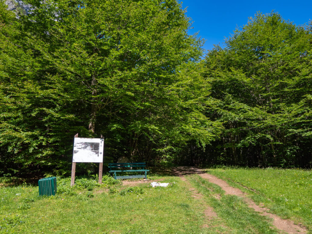 A faded sign and green bench in front of a forest marking the trailhead for the Three Peaks hike in Prokletije National Park.