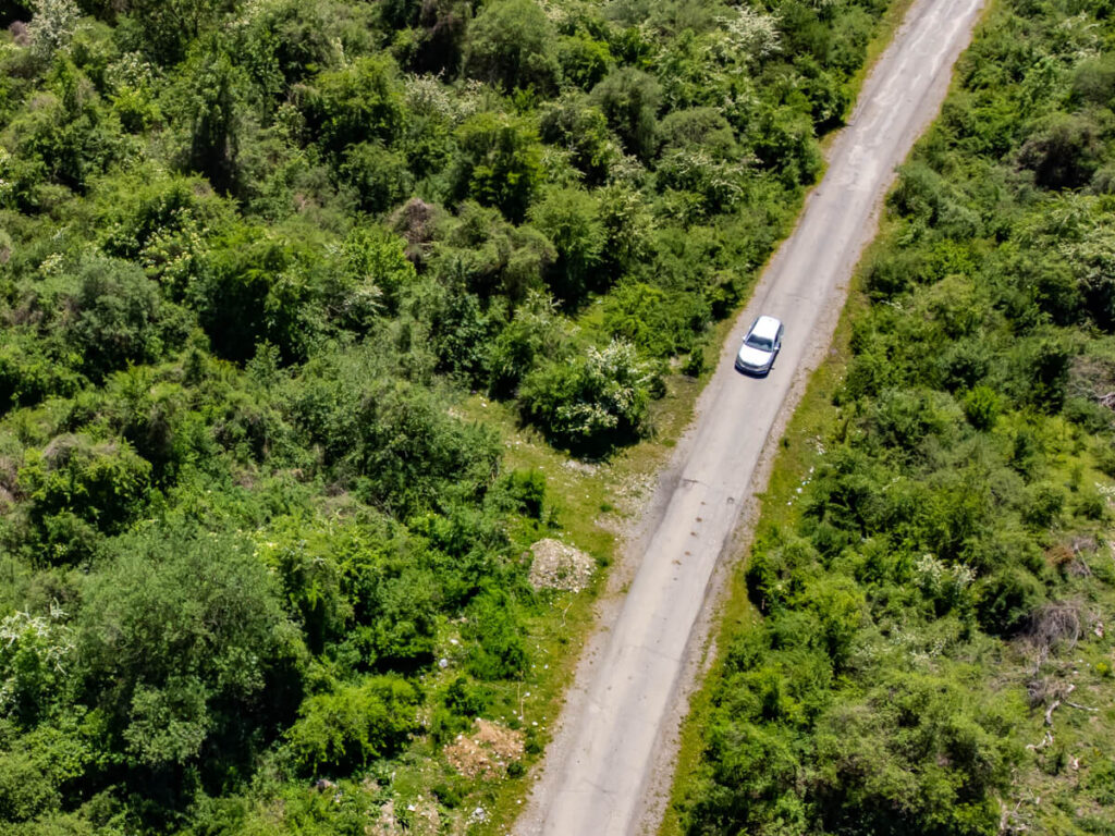 A drone view of a car driving down a single-track road surrounded by lush greenery in Prokletije National Park.