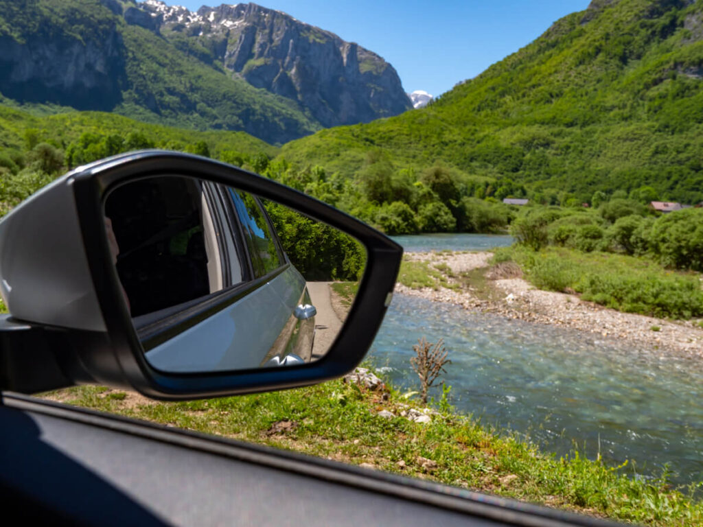 A car's wingmirror in front of a blue river running through the Ropojana Valley in Prokletije National Park.