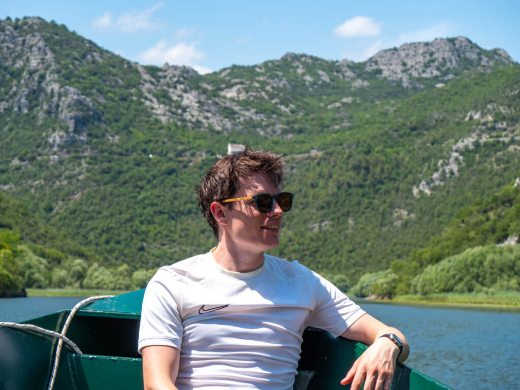 Dan smiling while sitting at the front of a boat on a river backed by the mountains of Lake Skadar.