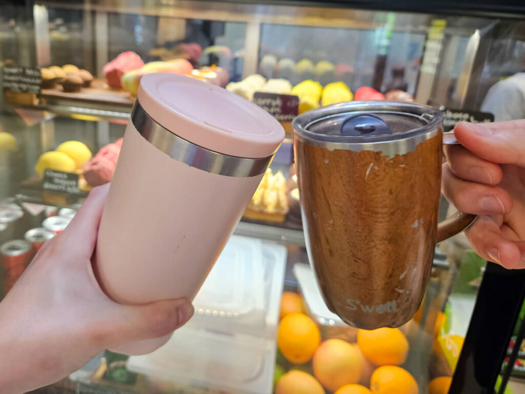 Two reusable coffee cups being cheersed together in front of a display of cakes at CAVE speciality coffee in Kotor.