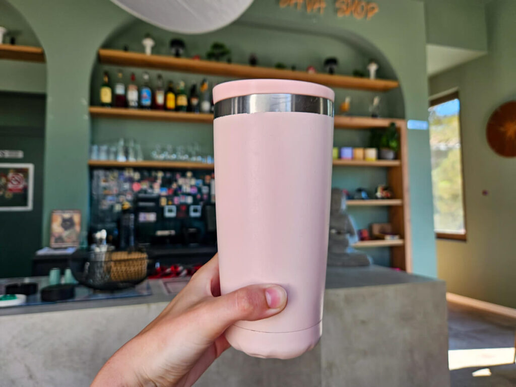 A pink reusable coffee cup held in front of a cafe counter in Java Coffee Centrale on the Luštica Peninsula.