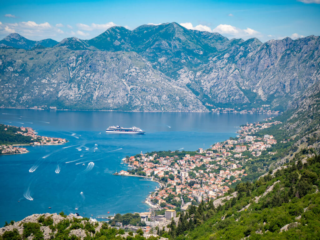 Neighbourhoods on the outskirts of Kotor lining the coast. A cruise ship sits in the Bay of Kotor, backed by mountains and surrounded by the white trails of speedboats.