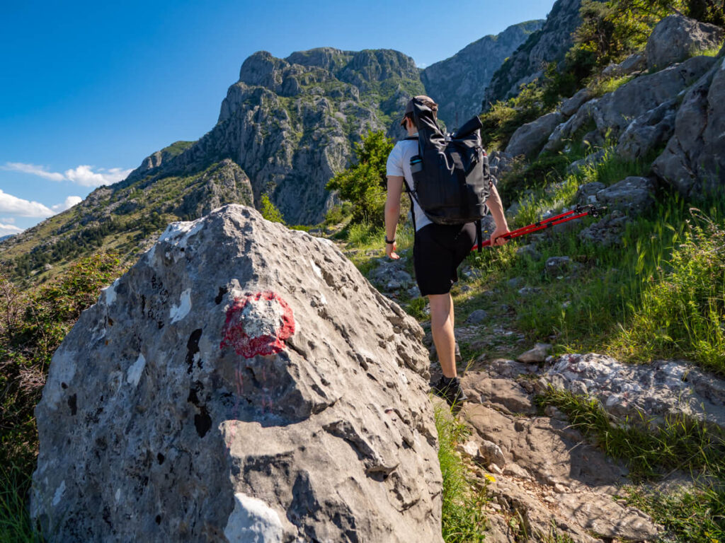 A red and white way marker painted on a rock with Dan climbing the Ladder of Kotor trail in the background.