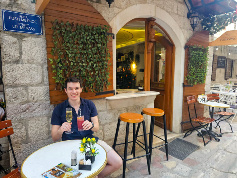 Dan sitting outside Patisserie by Wine House on the stone streets of Kotor holding two cocktails.