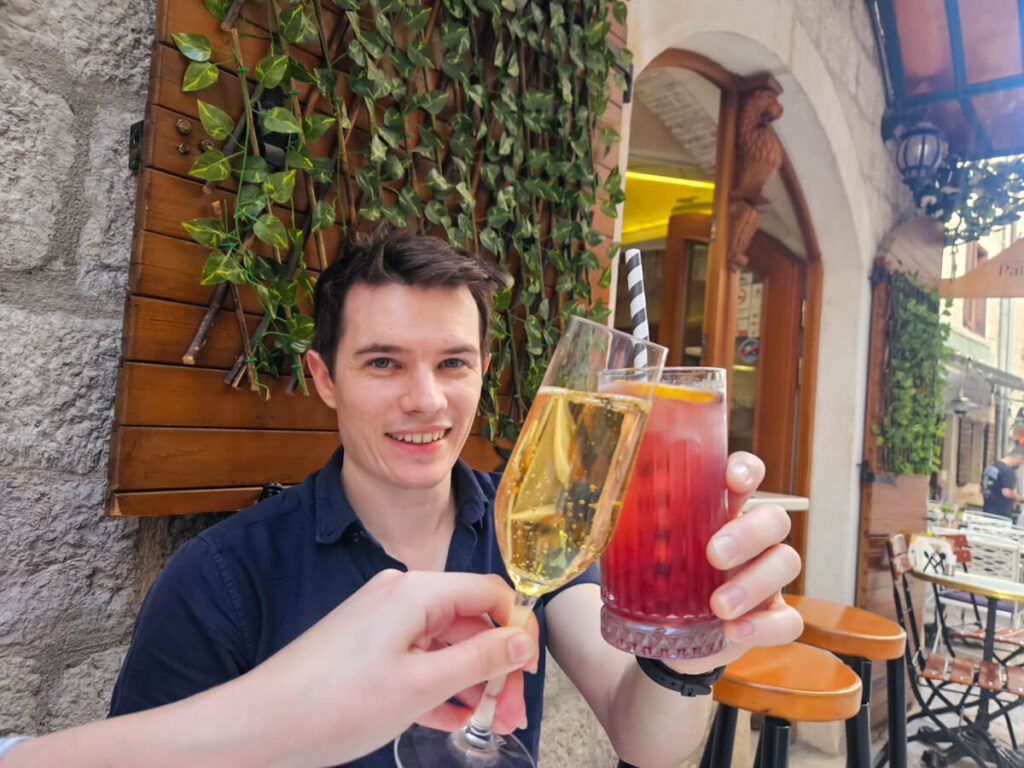 Dan cheersing his cocktail with a glass of prosecco at an outdoor table outside Patisserie by Wine House in Kotor.