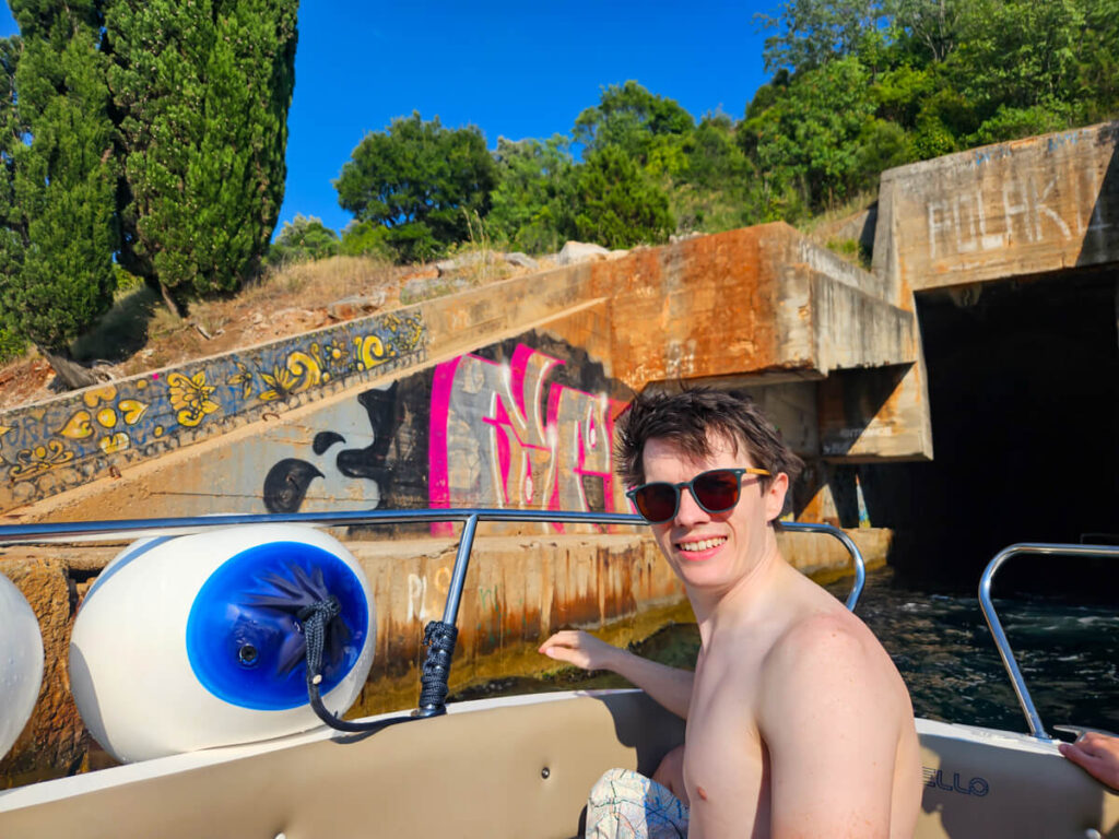 Dan smiling on a speedboat on a Blue Cave tour in front of the graffitied entrance to a submarine tunnel.