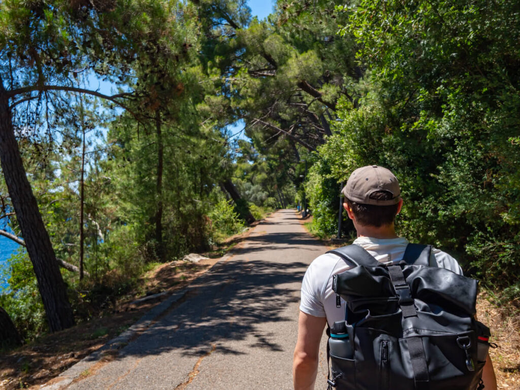 Dan walking along a tree-lined path along the headland near Petrovac, while wearing a backpack with two water bottles int he pockets.