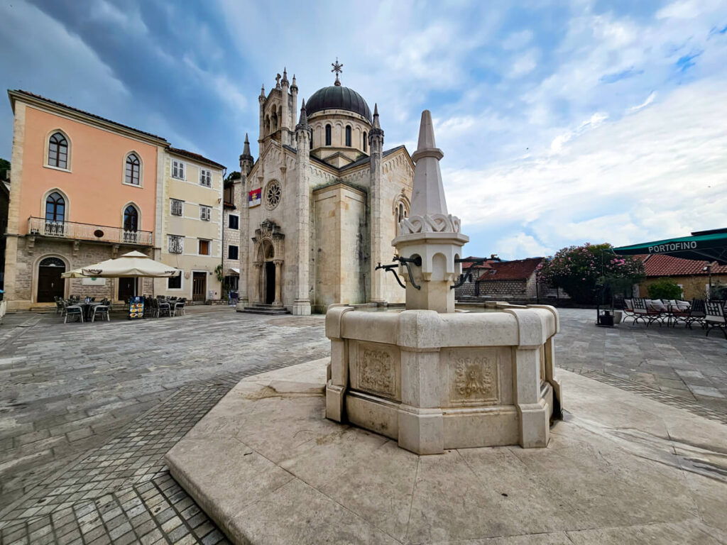 The main square in Herceg Novi's Old Town, complete with restaurants, a church, and a stone water fountain.