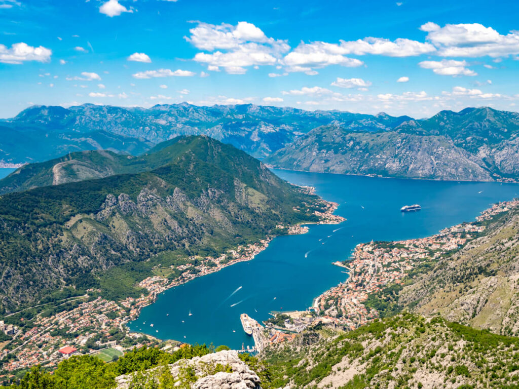 The blue waters of the Bay of Kotor lined by towns and surrounded by tall green mountains, viewed from the road to Lovćen National Park.