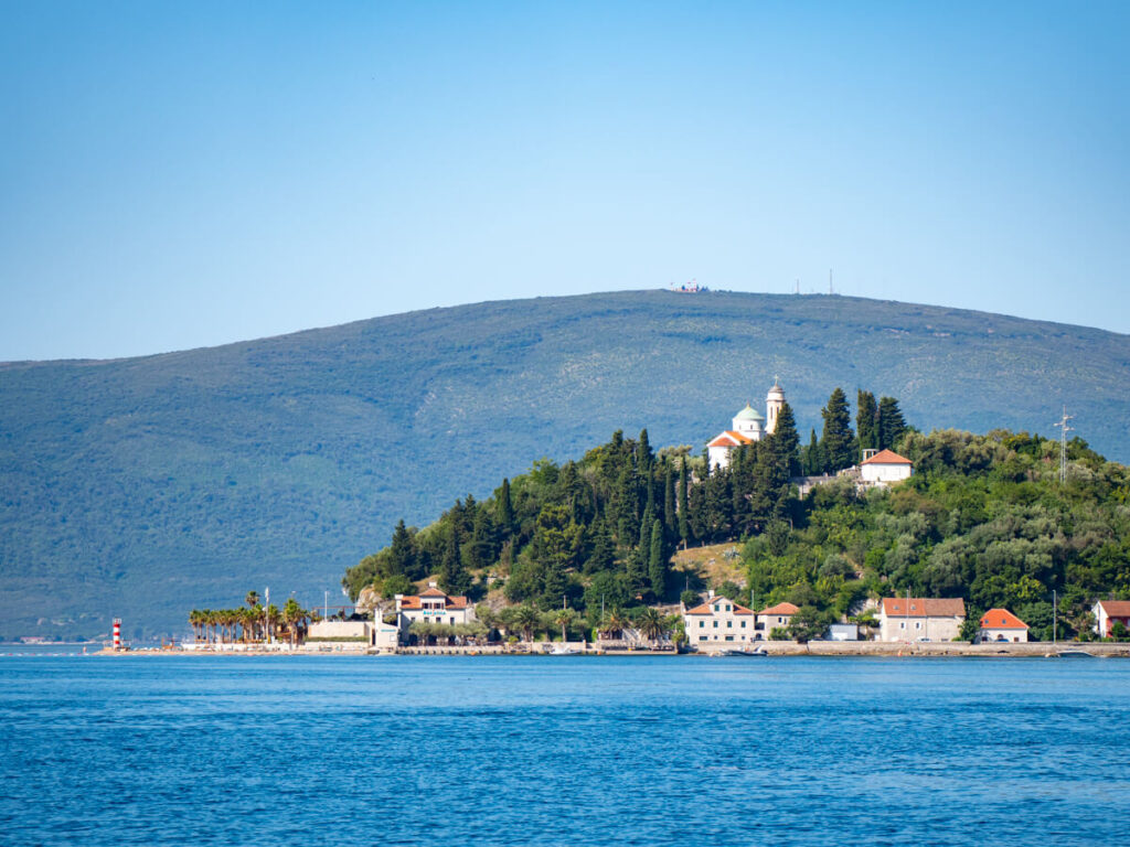 A small dome-shaped island in the Bay of Kotor with trees and some small buildings.