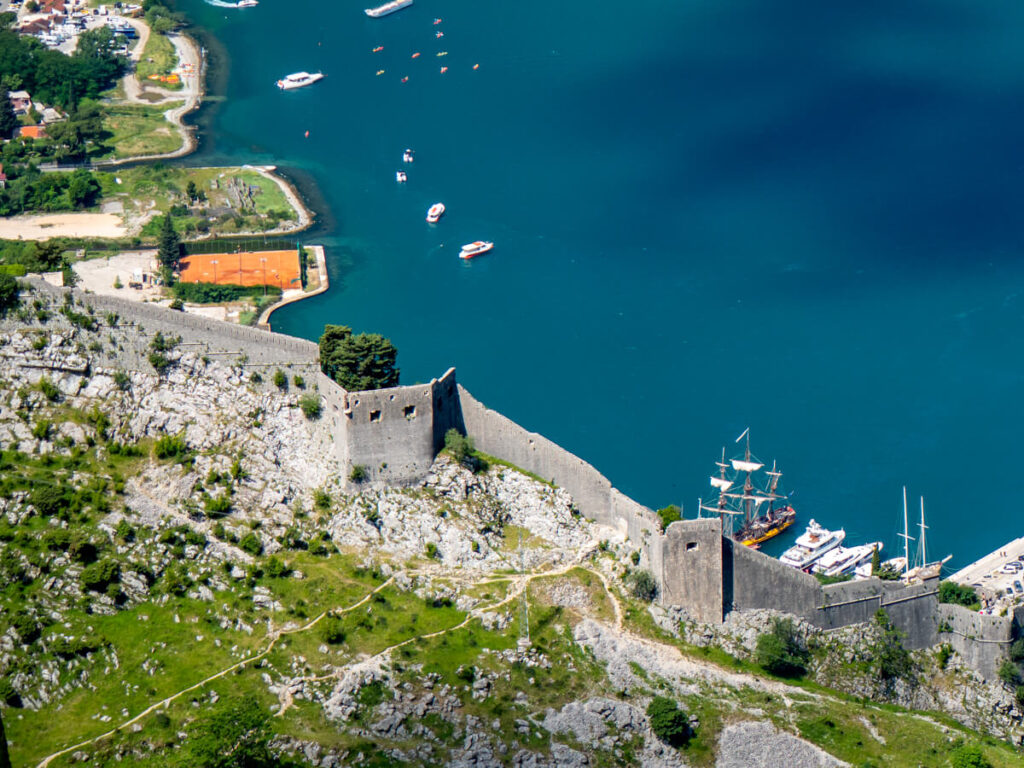 The stone wall of the medieval San Giovanni Fortress snaking down a hill and backed by bright blue waters of the Bay of Kotor.