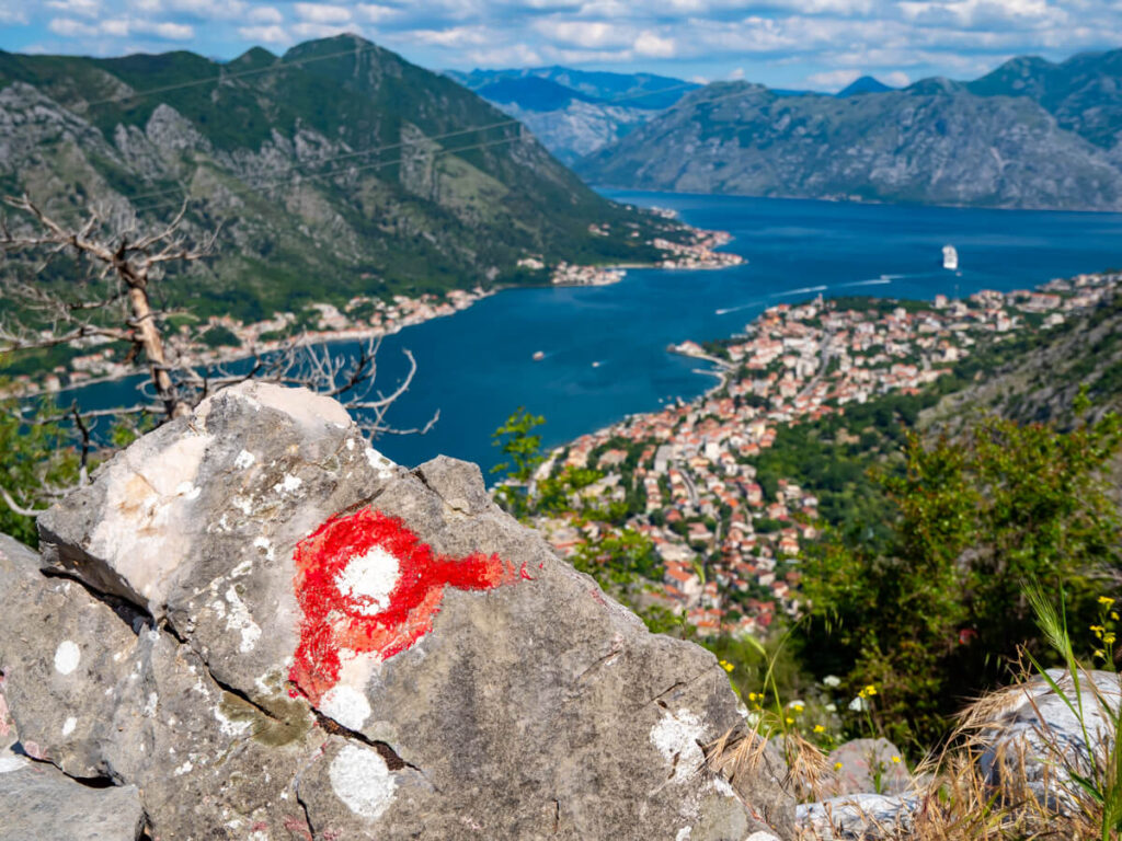 A red and white trail marker painted on a rock above the blue waters of the Bay of Kotor.