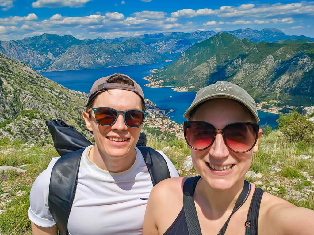 Lucy and Dan in hats and sunglasses smiling in front of a view of the Bay of Kotor.