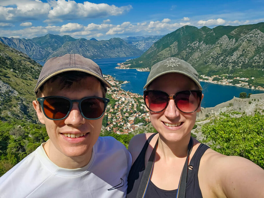 Lucy and Dan smiling in front of the blue Bay of Kotor at the top of the Ladder of Kotor.