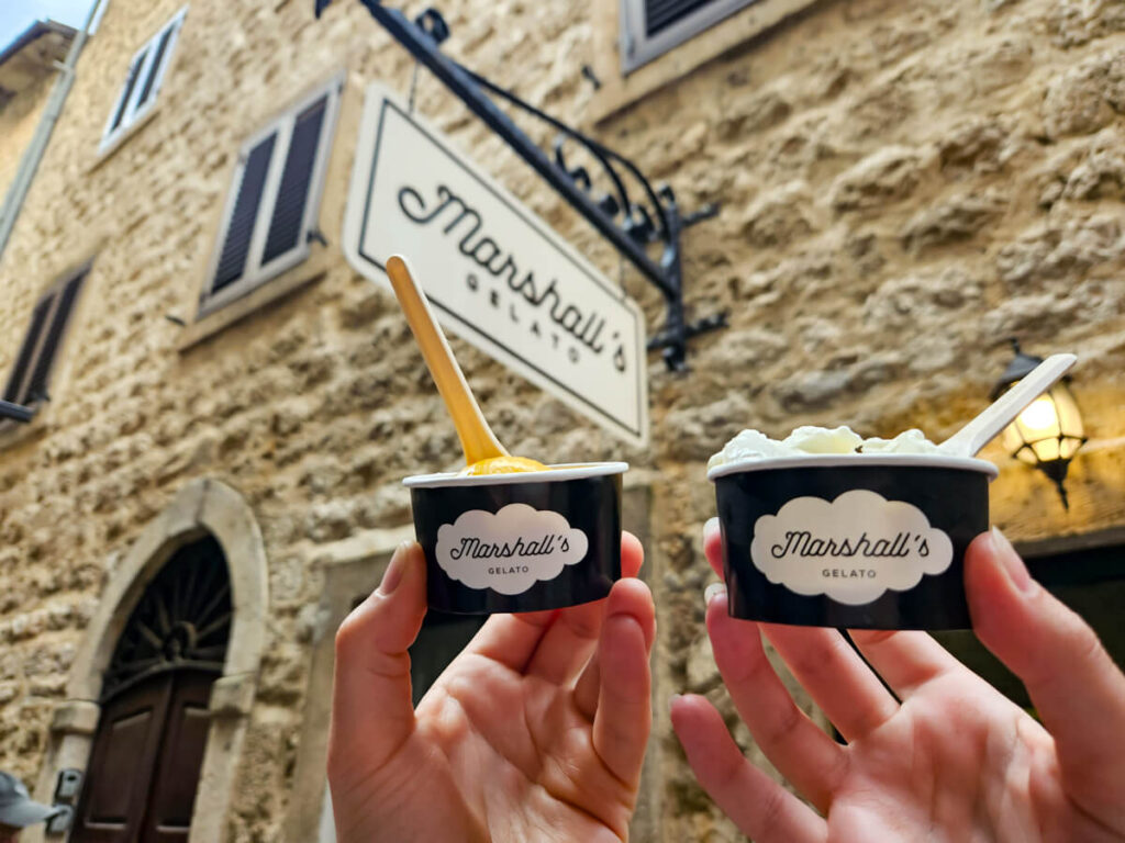 Two pots of gelato held in front of a sign for Marshall's Gelato in Kotor.