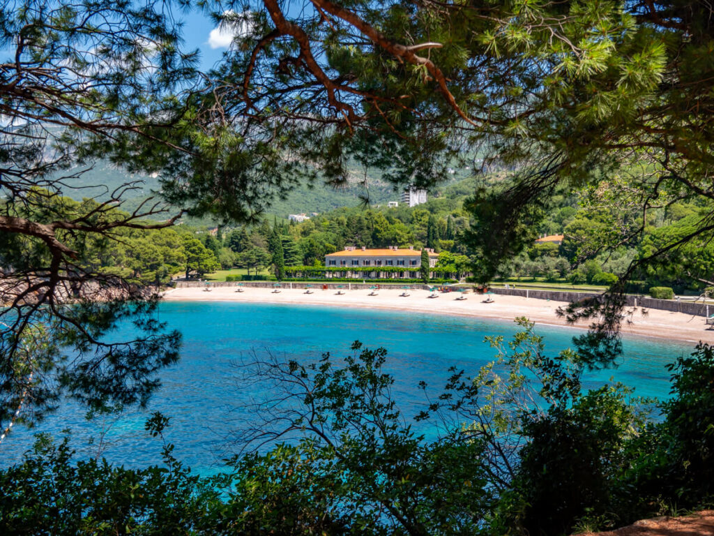 The curved white sand and turquoise waters of Miločer Beach peeking through the branches of a park.