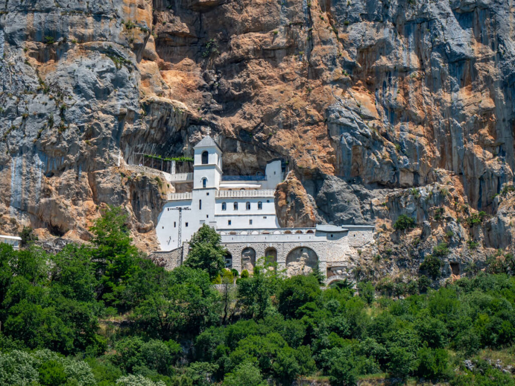 The whitewashed façade of the upper part of Ostrog Monastery caved into a cliff and surrounded by greenery.