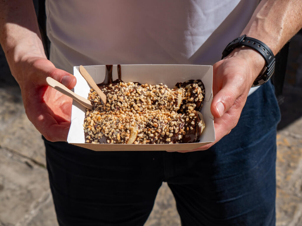Dan's hands holding a paper tray of small fluffy pancakes covered in chocolate sauce and nuts in Budva.