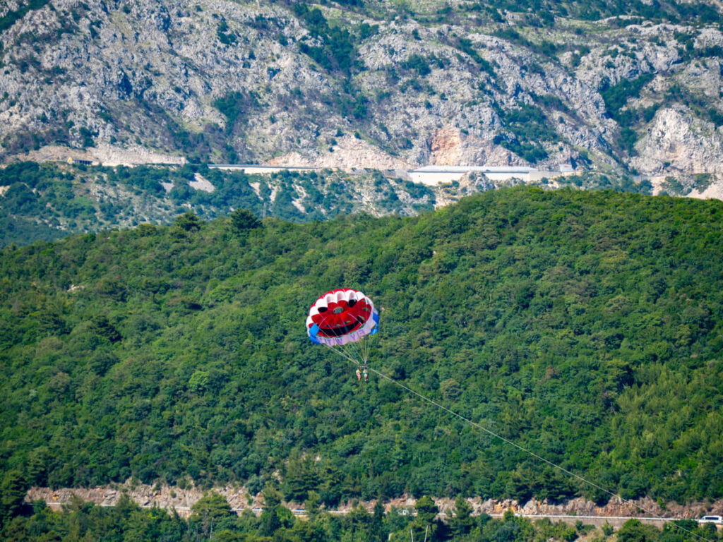A couple parasailing in Budva, the red balloon against a green mountain backdrop.