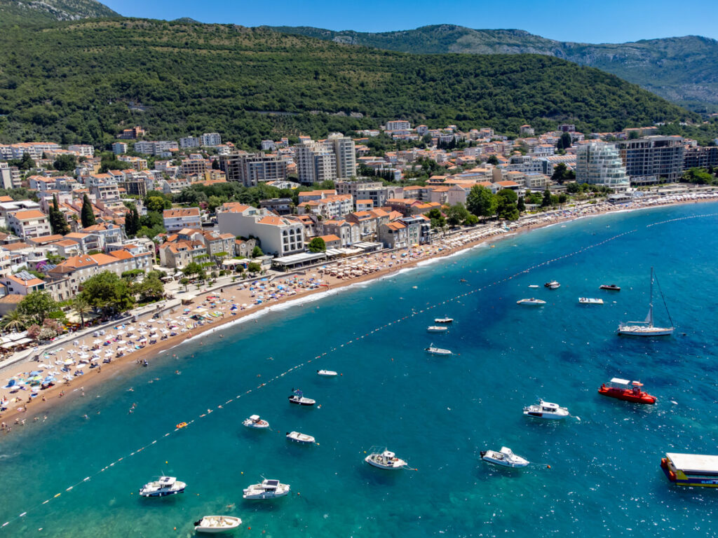 A drone capture of boats bobbing on the turquoise seas by a long sandy beach covered in umbrellas lined by the coastal town of Petrovac.