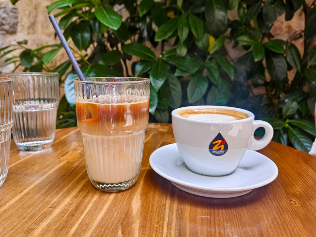 Two coffees, one hot, one ices, on a table in a garden with plants at Piccolo Café in Kotor.