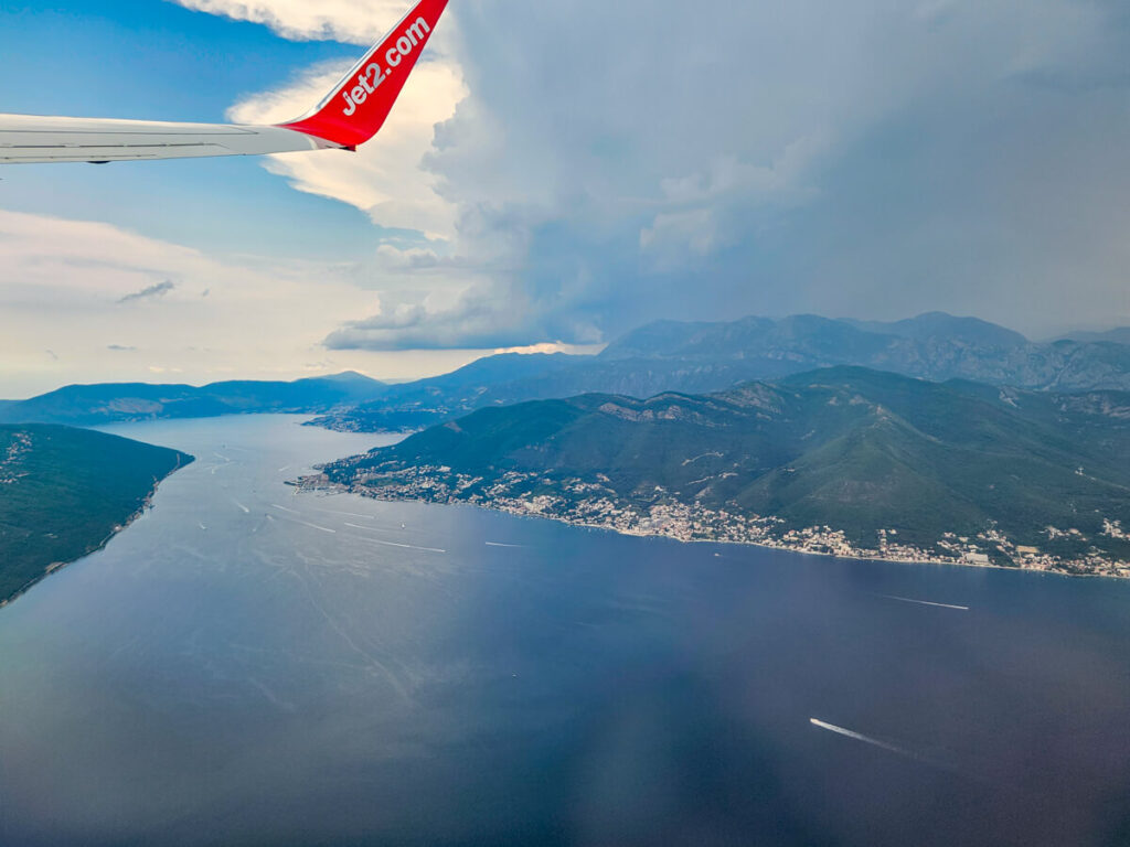 A Jet2.com plane wing flying over the Bay of Kotor. Clouds are beginning to roll on and cover the once blue sky.