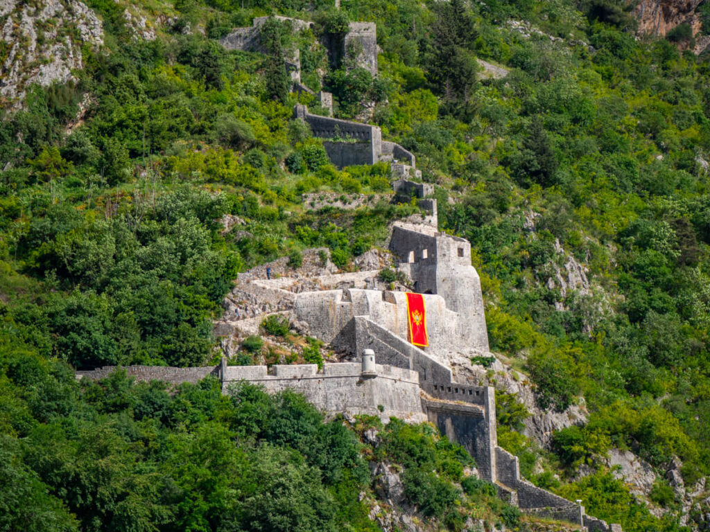 A winding stone wall leading down a green hill to the San Giovanni Fortress in Kotor.