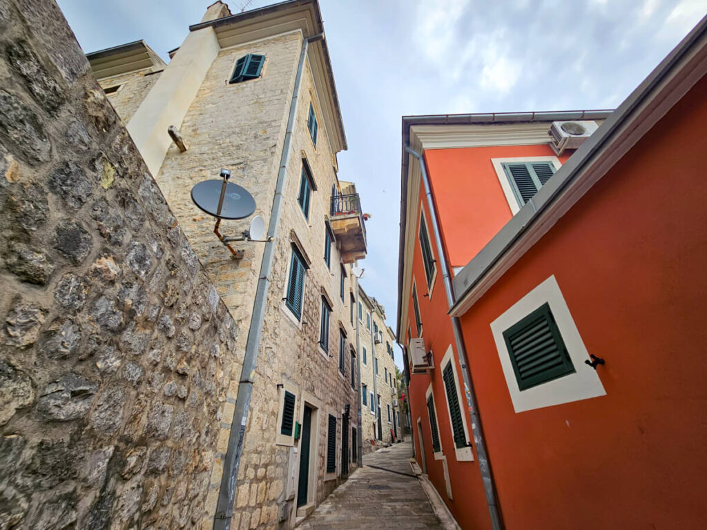 A tight cobbled street in Herceg Novi with stone buildings on one side, and brightly-painted houses on the other.