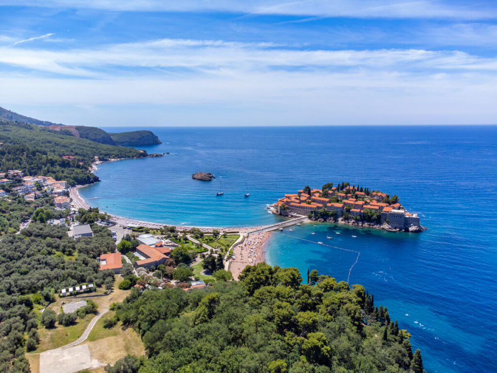 A drone view of Sveti Stefan with the causeway leading to an island of red-rooved houses and a lush park lining sandy beaches.