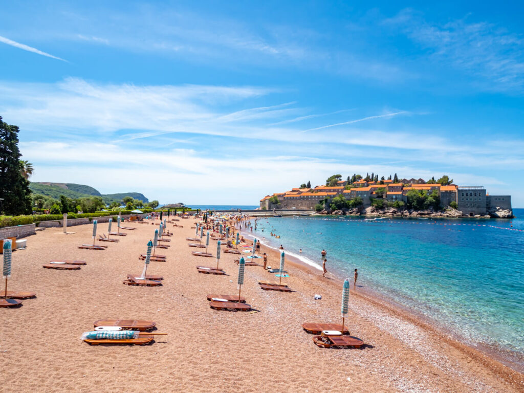 Beach umbrellas on a pink-pebbled beach leading to the iconic Sveti Stefan Island.
