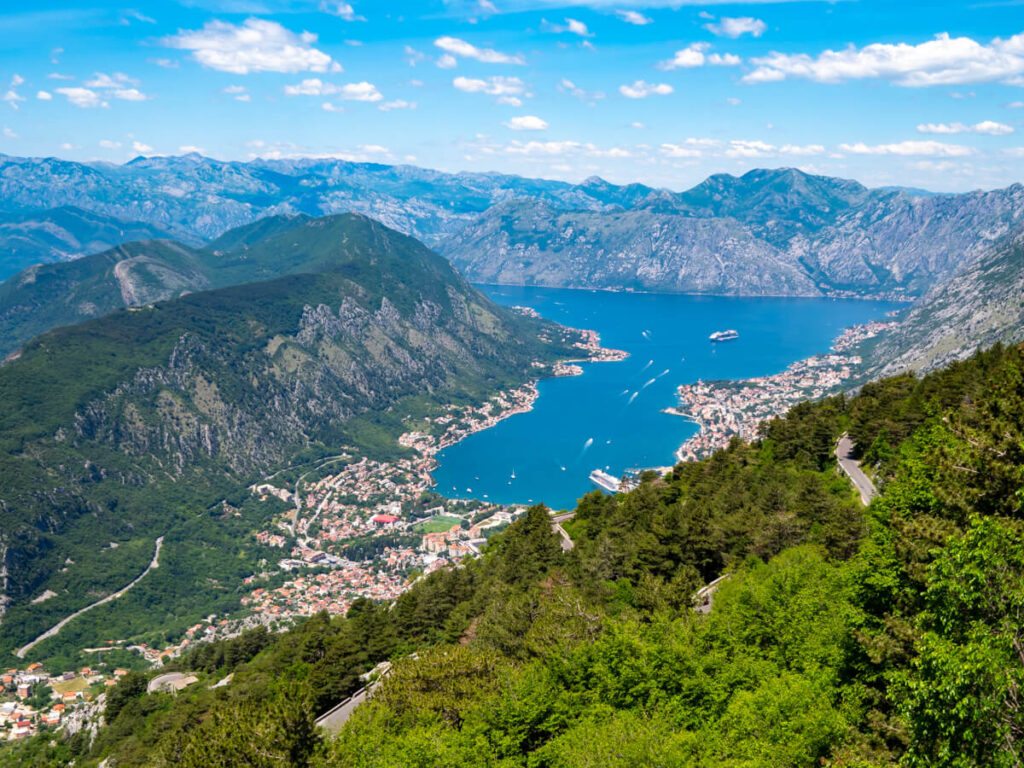 A road on a mountainside backed by the blue waters of the Bay of Kotor and surrounded by green mountains and lined with small towns.