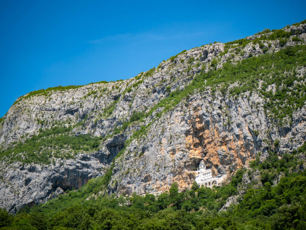 The small whitewashed façade of Ostrog Monastery carved into a huge limestone cliff surrounded by greenery.