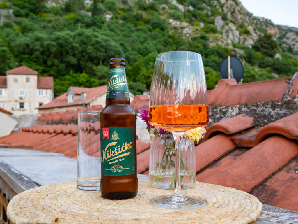 A glass of rose wine and a bottle of Niksicko beer on a table at a rooftop restaurant in Kotor backed by the mountains.